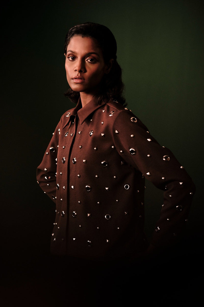 ROSANI The Monolith jacket in brown - collared long-sleeve jacket with all-over metal stud and ring embellishments, editorial three-quarter view on dark green background