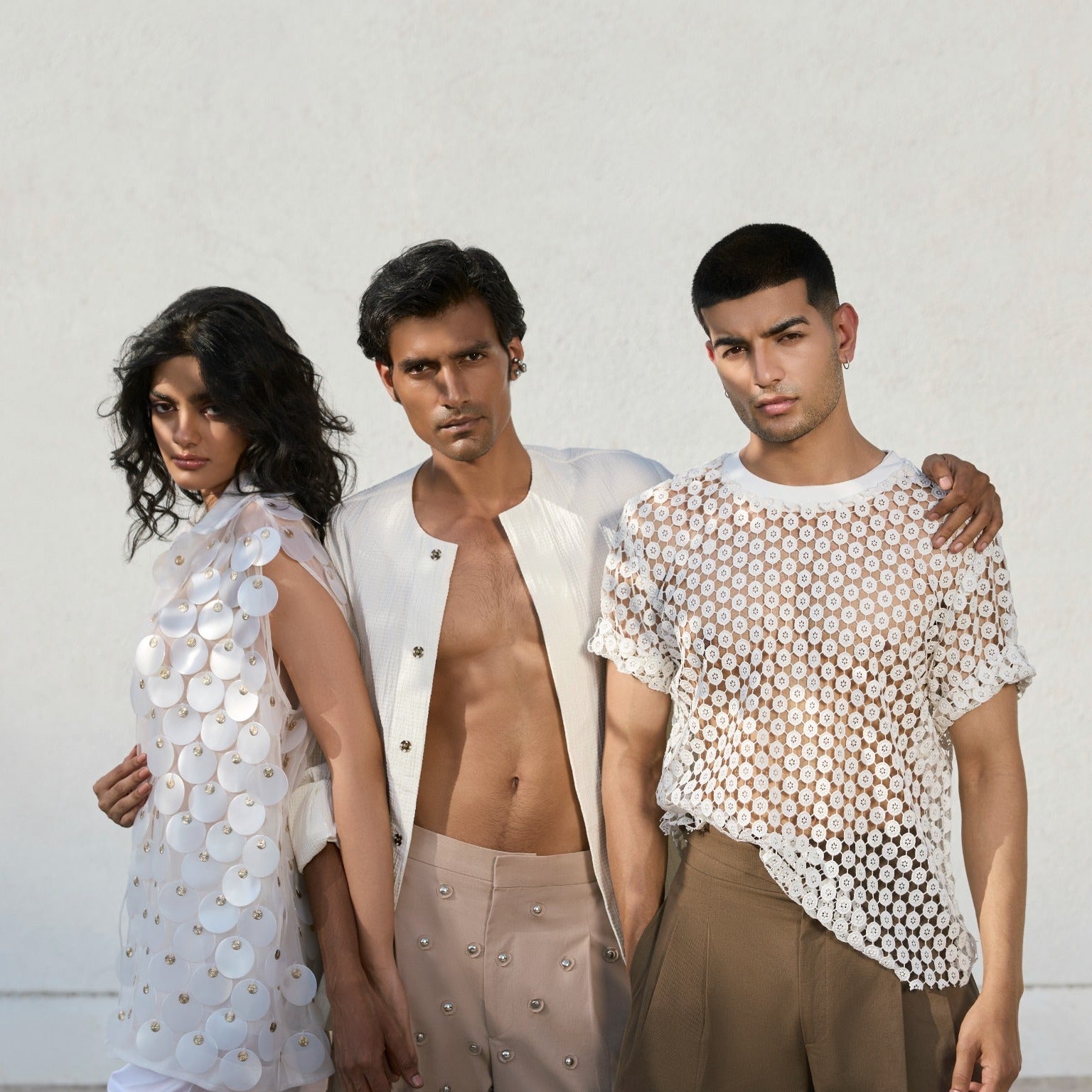 Three models in a studio group portrait wearing ROSANI CONVERGENCE looks: woman in white disc-embellished vest dress, man in cream sleeveless vest with nude studded trousers, and man in white lace t-shirt with brown trousers