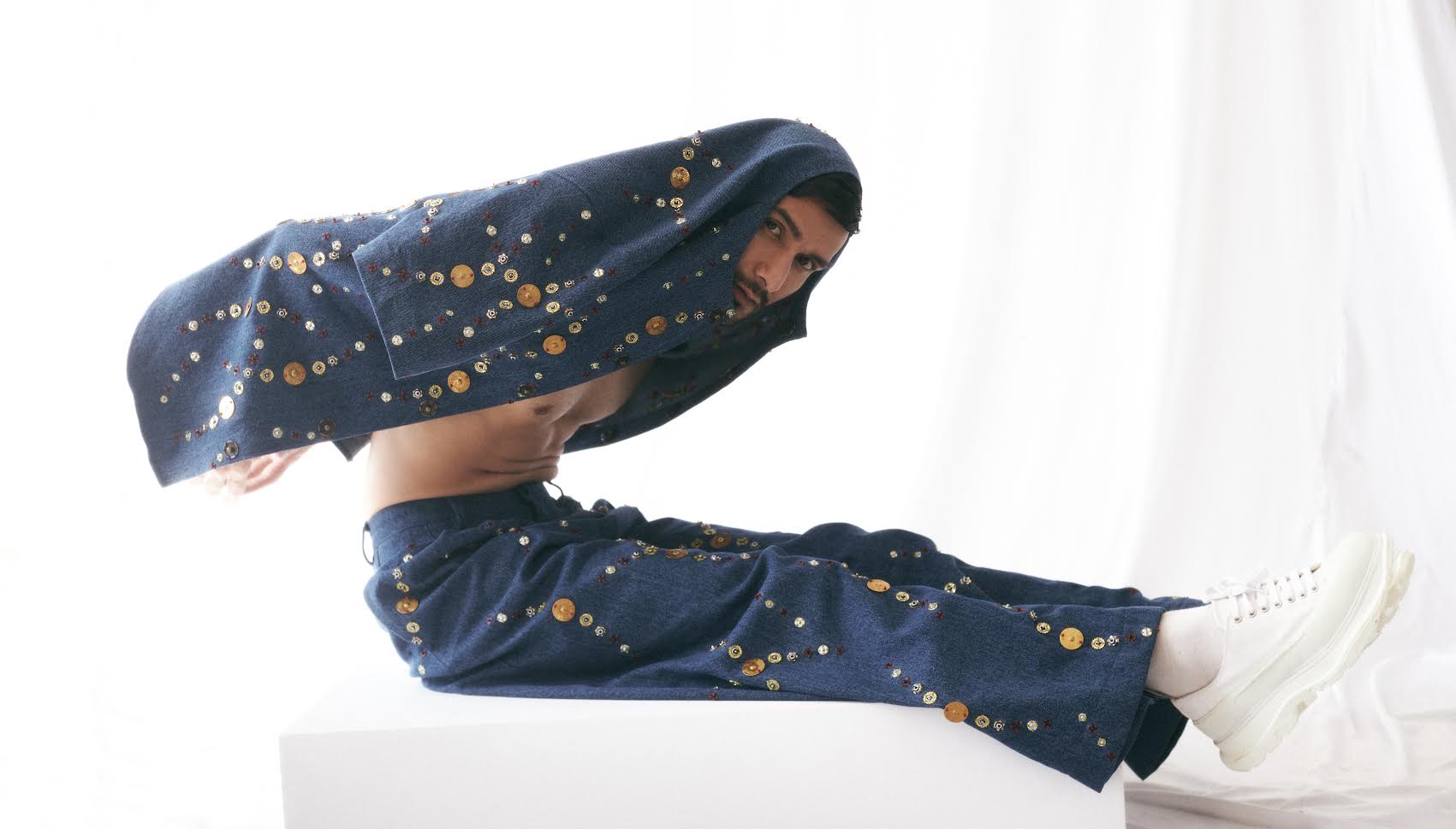 ROSANI DYMENSION collection image - model wearing The Atto shirt and The Enzo jeans in midnight denim with metallic button embellishment, reclining pose on white set, front view